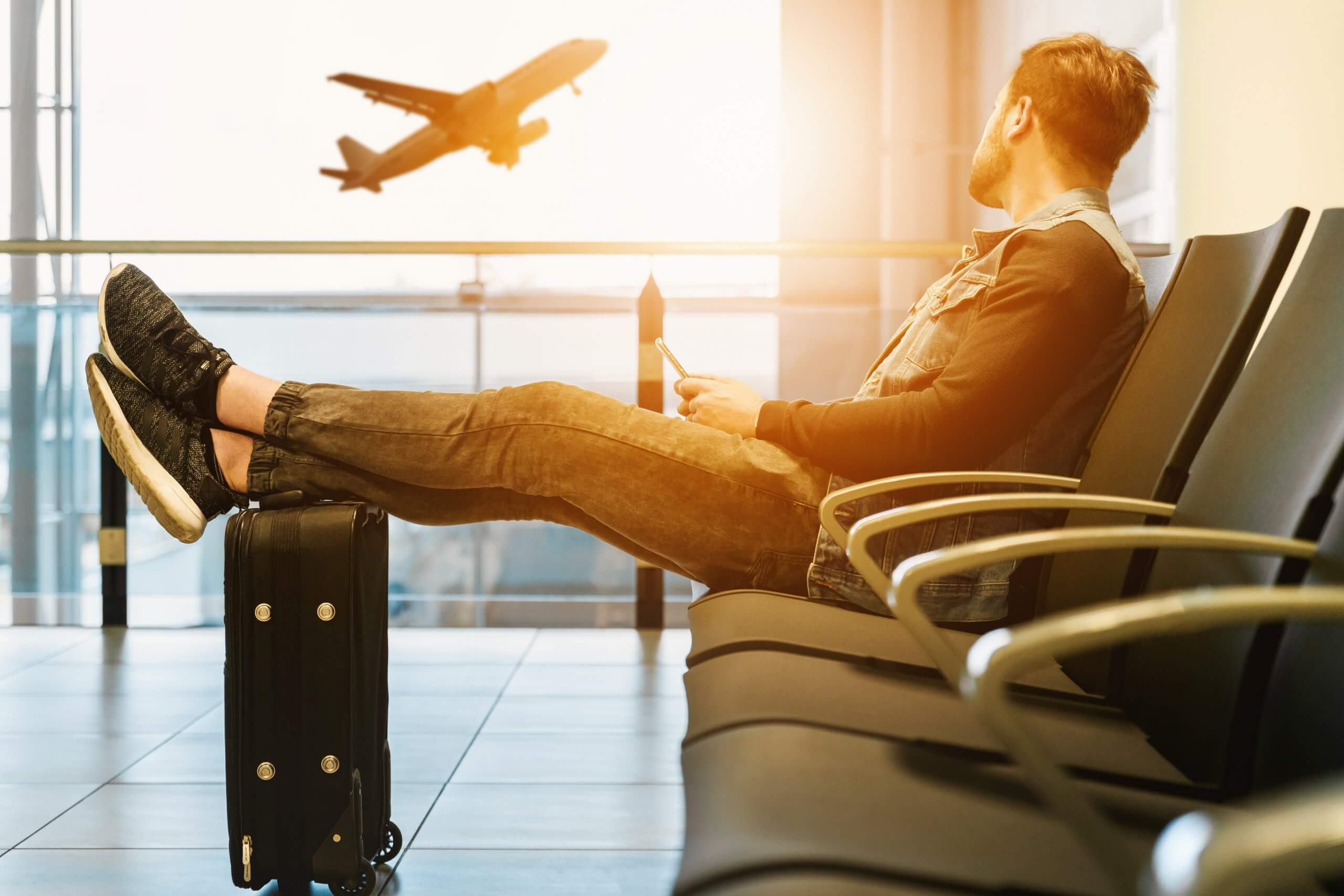 man in airport with legs up on his hand luggage while watching a plane take off outside the window