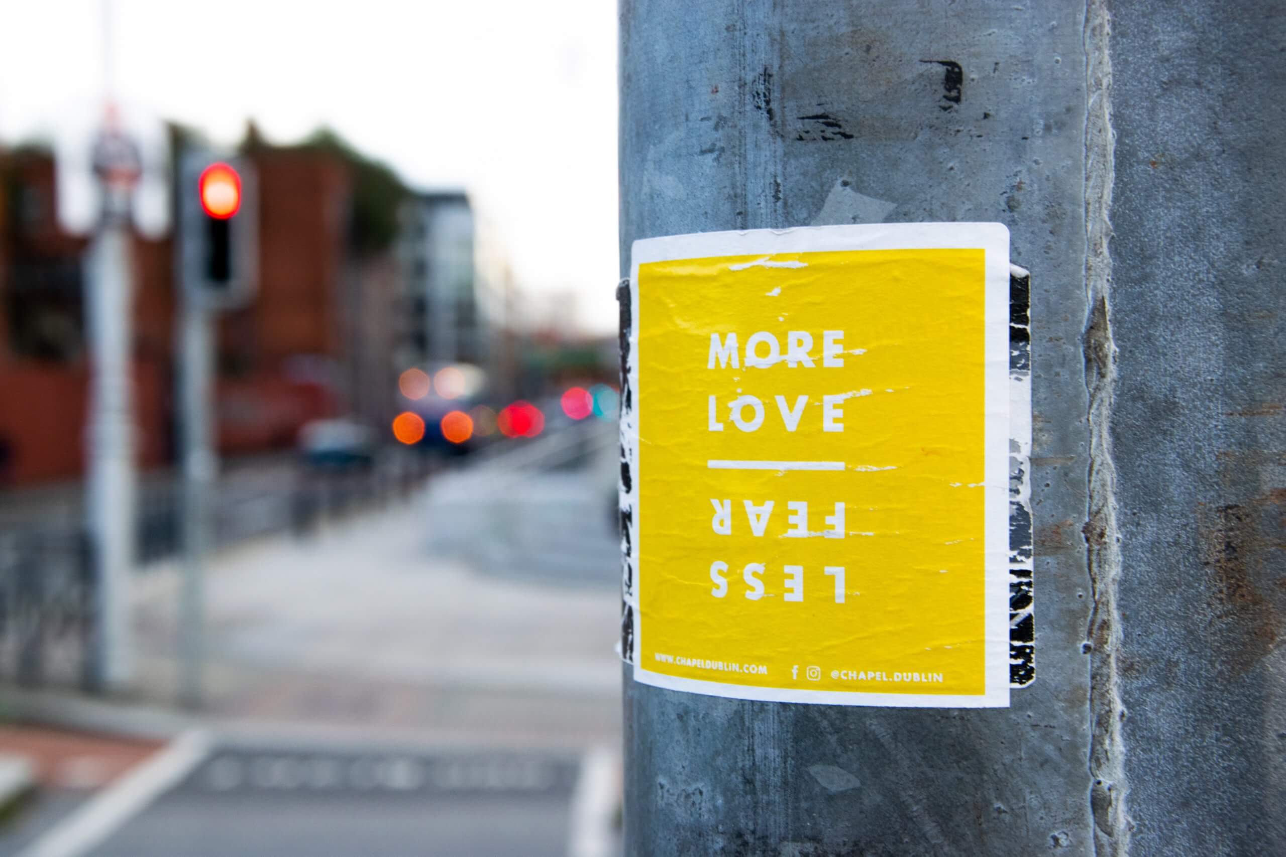 a lamp post in focus against a blurred street scene with a yellow sign declaring "more love, less fear"
