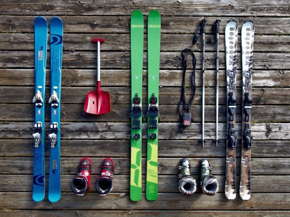 three sets of skis in blue, green and white lying on a wooden decking with boots, poles and a shovel around them