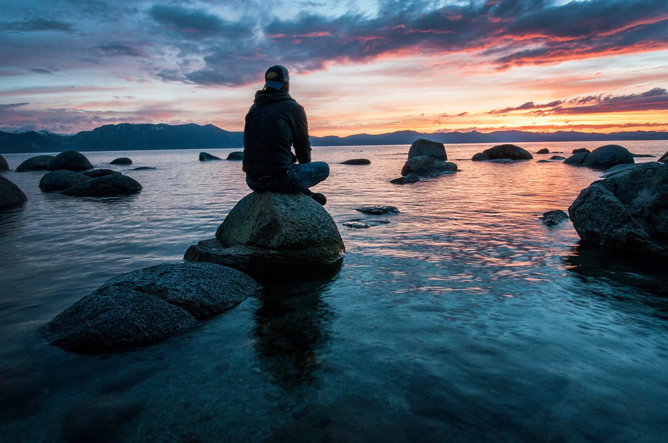 man sitting crosslegged on a rock in the middle of a lake looking out at a beautiful sunset
