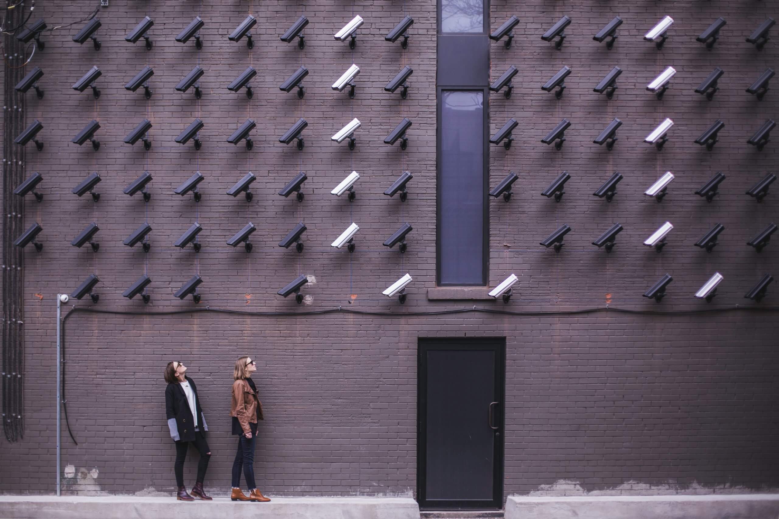 two people looking up at a sea of surveillance cameras mounted on brick wall
